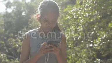 亚洲可爱的女孩使用手机带耳机，早上站在红<strong>树林阳光下</strong>。